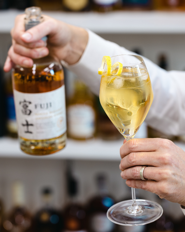 a person holding a bottle of whisky and a wine glass in front of a whisky shelf