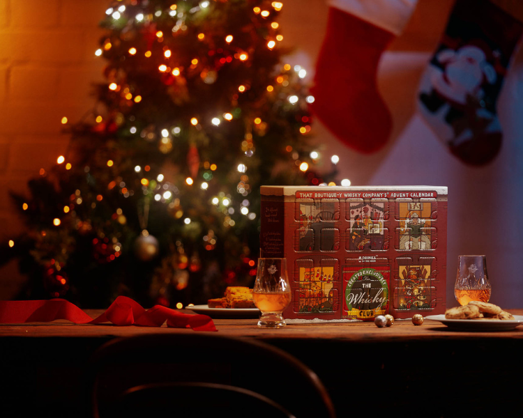 The Whisky Club's 2021 Advent Calendar on a table  with cookies and whiskies and a Christmas tree with lights in the background