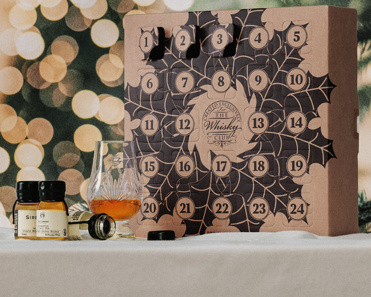 A whisky advent calendar and a Glencairn of whisky on a white tablecloth with a Christmas tree in the background 